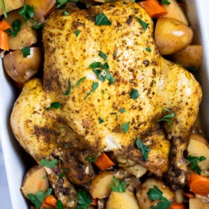 A whole chicken, broiled until crispy, resting on top of potatoes and carrots in a baking dish, topped with fresh parsley.