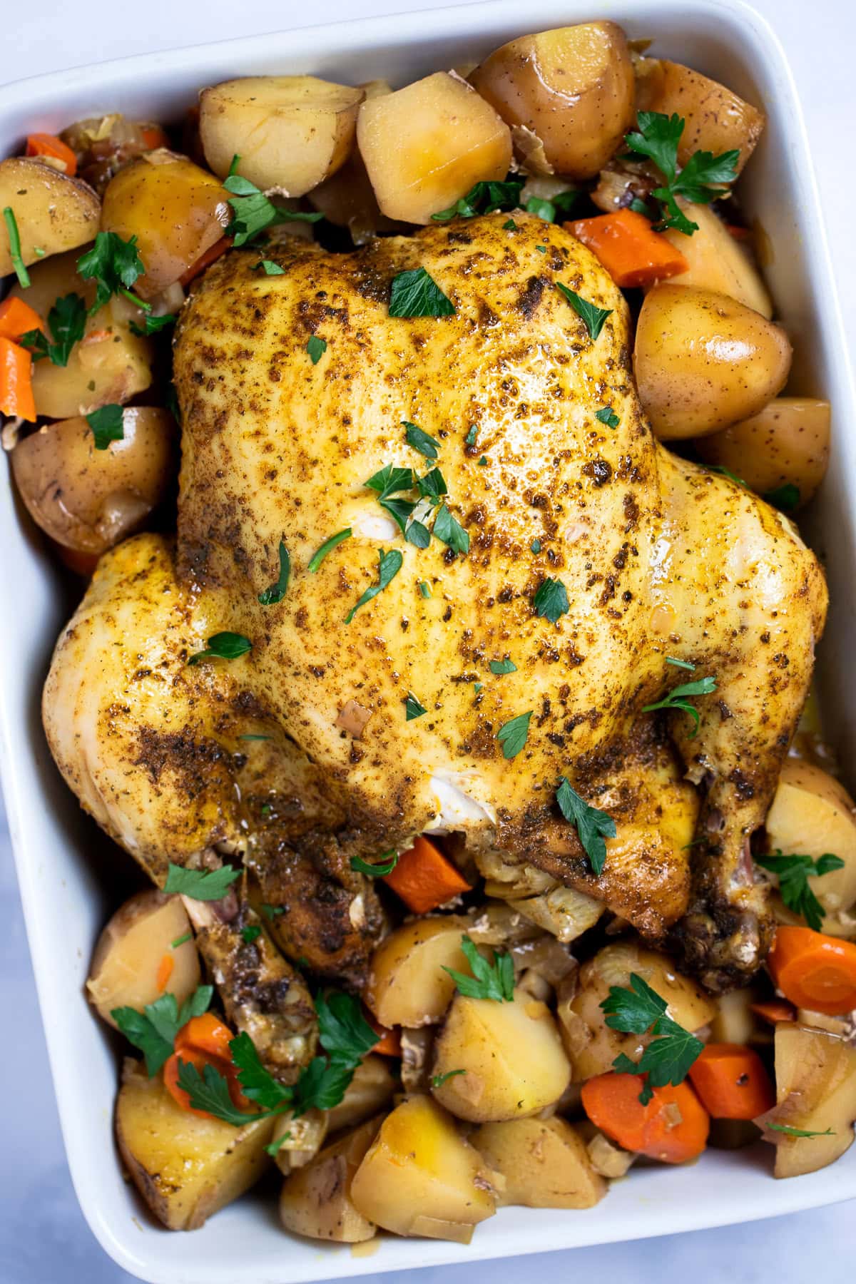 A whole chicken, broiled until crispy, resting on top of potatoes and carrots in a baking dish, topped with fresh parsley.