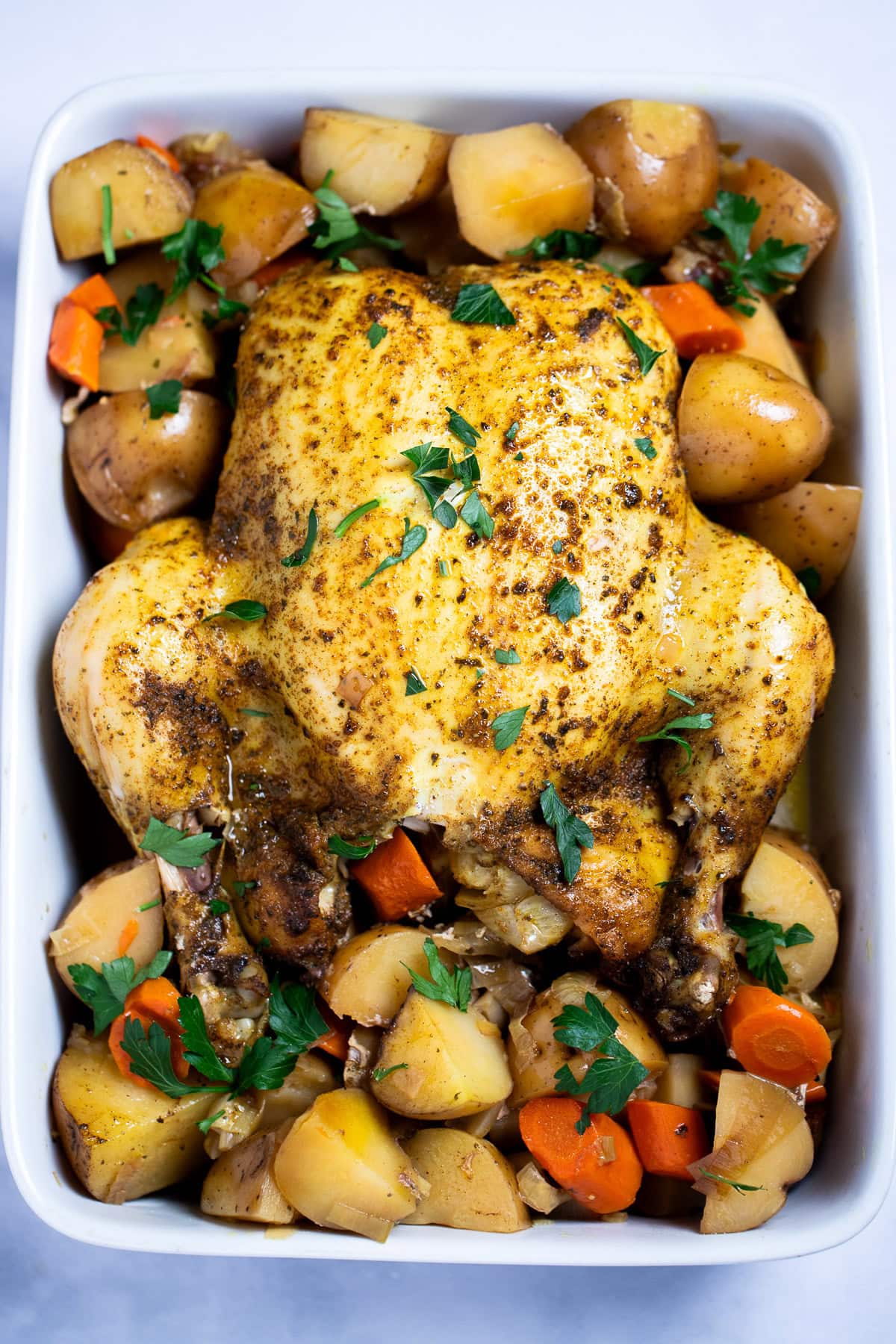 A whole chicken, broiled until crispy, resting on top of potatoes and carrots in a baking dish, topped with fresh parsley.