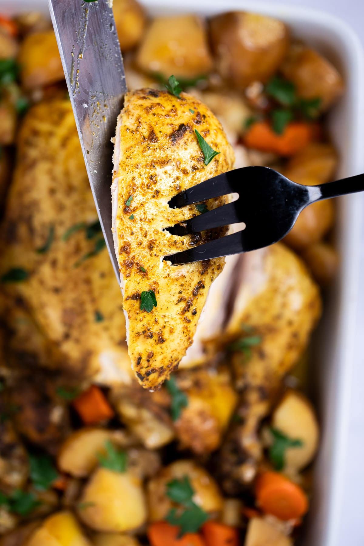 A knife and fork holding a chicken breast up after carving it from a whole chicken sitting in a baking dish with potatoes and fresh parsley.