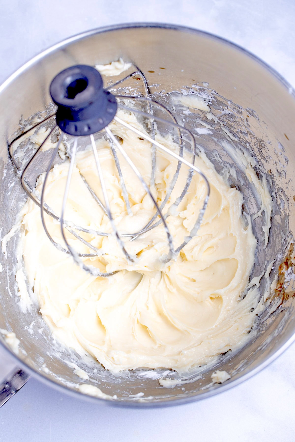 A mixing bowl with creamed cream cheese on a table with a stand mixer whisk in it.