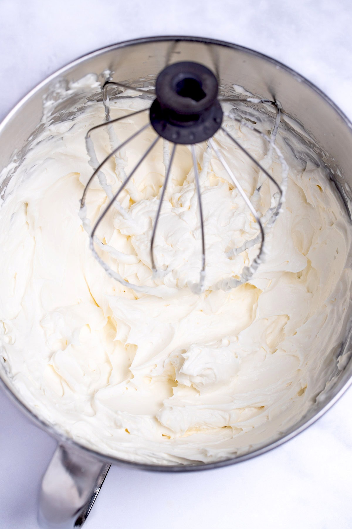 A mixing bowl with whipped cream on a table, with a stand mixer whisk in it.