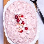 A serving bowl with cranberry fluff salad topped with mini marshmallows and fresh cranberries, resting on a cutting board, next to a cup of fresh cranberries and a serving spoon.