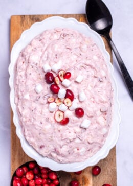 A serving bowl with cranberry fluff salad topped with mini marshmallows and fresh cranberries, resting on a cutting board, next to a cup of fresh cranberries and a serving spoon.