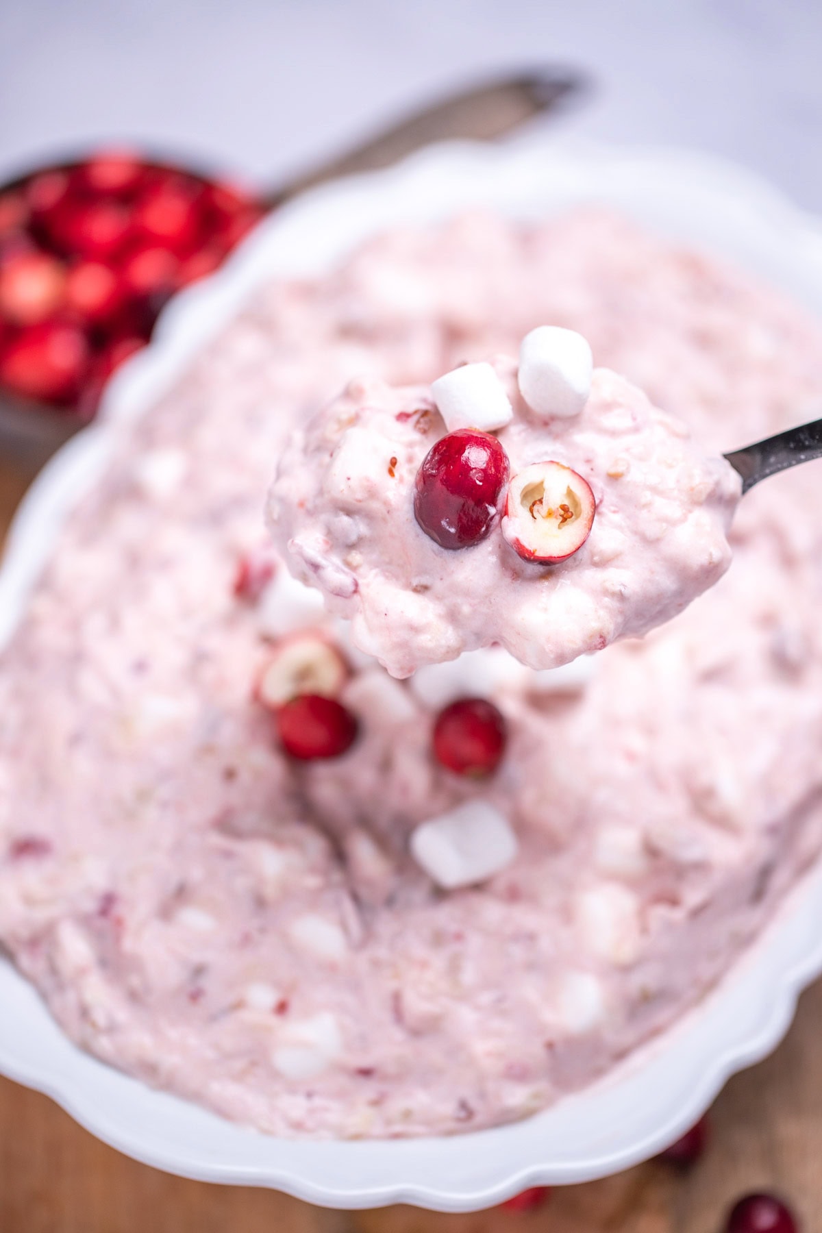 A serving bowl with cranberry fluff salad topped with mini marshmallows and fresh cranberries, resting on a cutting board, next to a cup of fresh cranberries and a serving spoon lifting some fluff up.