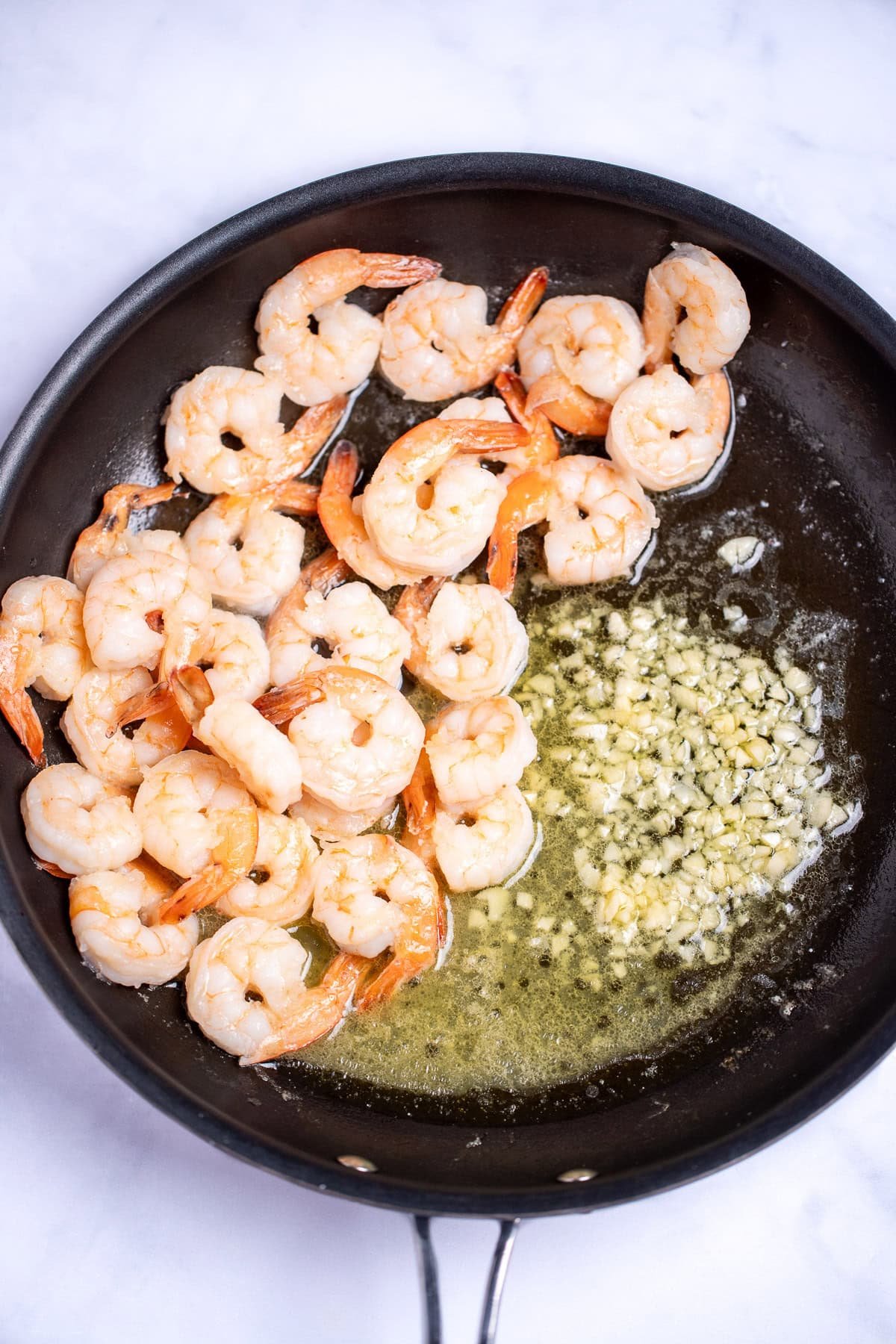 A nonstick skillet with shrimp pushed to one side, and minced garlic cooking in butter on the other side.