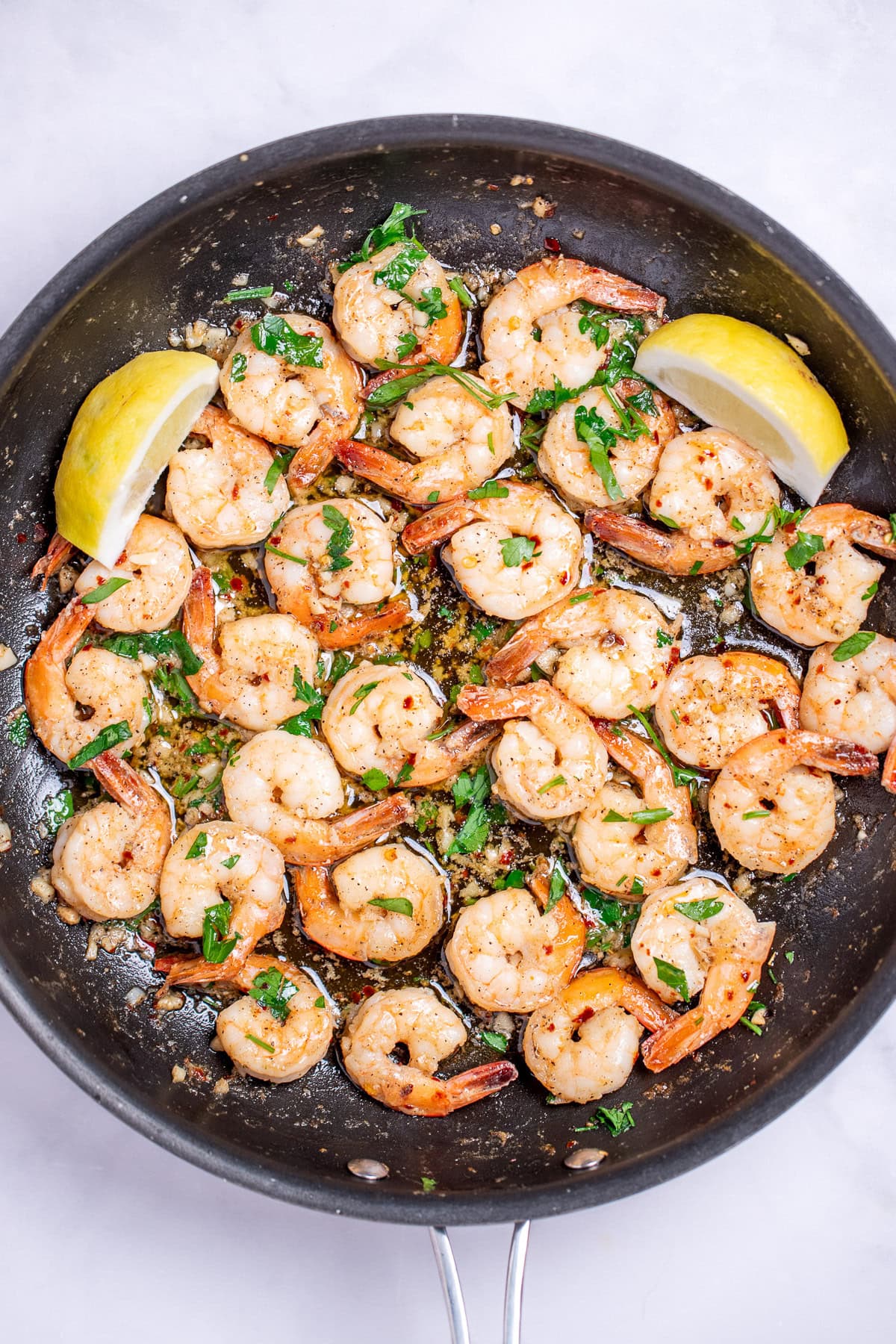A nonstick skillet with cooked shrimp in garlic and butter with fresh parsley and two lemon wedges in the skillet.