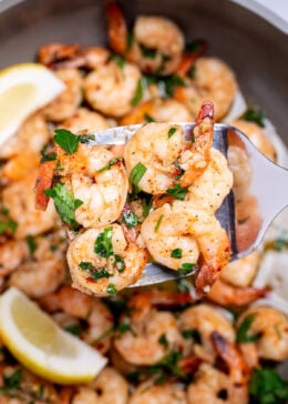 A nonstick skillet with cooked shrimp in garlic and butter with fresh parsley and two lemon wedges in the skillet, with a spatula lifting some up.