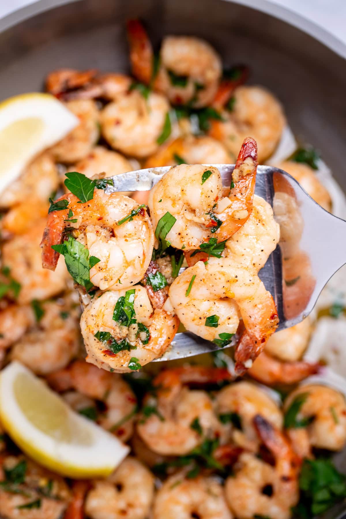 A nonstick skillet with cooked shrimp in garlic and butter with fresh parsley and two lemon wedges in the skillet, with a spatula lifting some up.