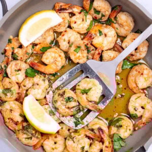 A nonstick skillet with cooked shrimp in garlic and butter with fresh parsley and two lemon wedges in the skillet, with a spatula holding some shrimp and resting in the pan.