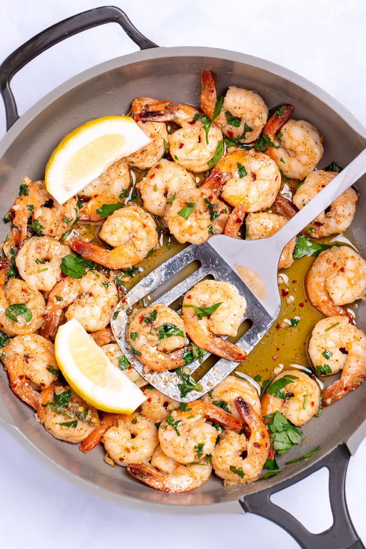 A nonstick skillet with cooked shrimp in garlic and butter with fresh parsley and two lemon wedges in the skillet, with a spatula holding some shrimp and resting in the pan.
