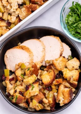 A bowl with gluten free stuffing and turkey slices on a table, in front of a baking dish of stuffing, and next to a small glass bowl of chopped fresh parsley.