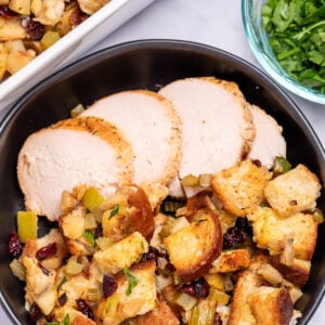 A bowl with gluten free stuffing and turkey slices on a table, in front of a baking dish of stuffing, and next to a small glass bowl of chopped fresh parsley.