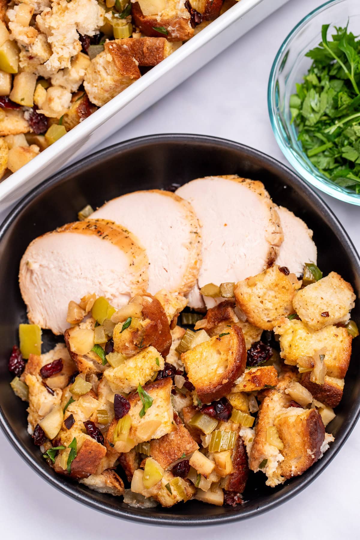 A bowl with gluten free stuffing and turkey slices on a table, in front of a baking dish of stuffing, and next to a small glass bowl of chopped fresh parsley.