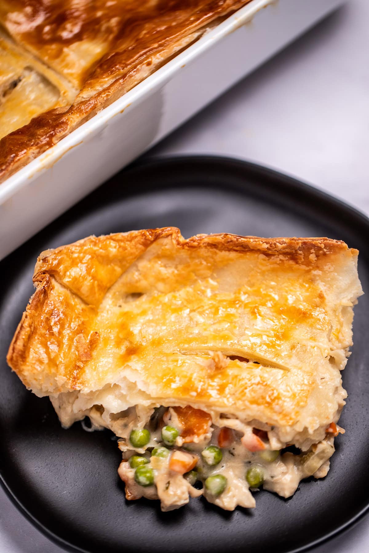 A plate with a piece of pot pie casserole on a table in front of a casserole dish.