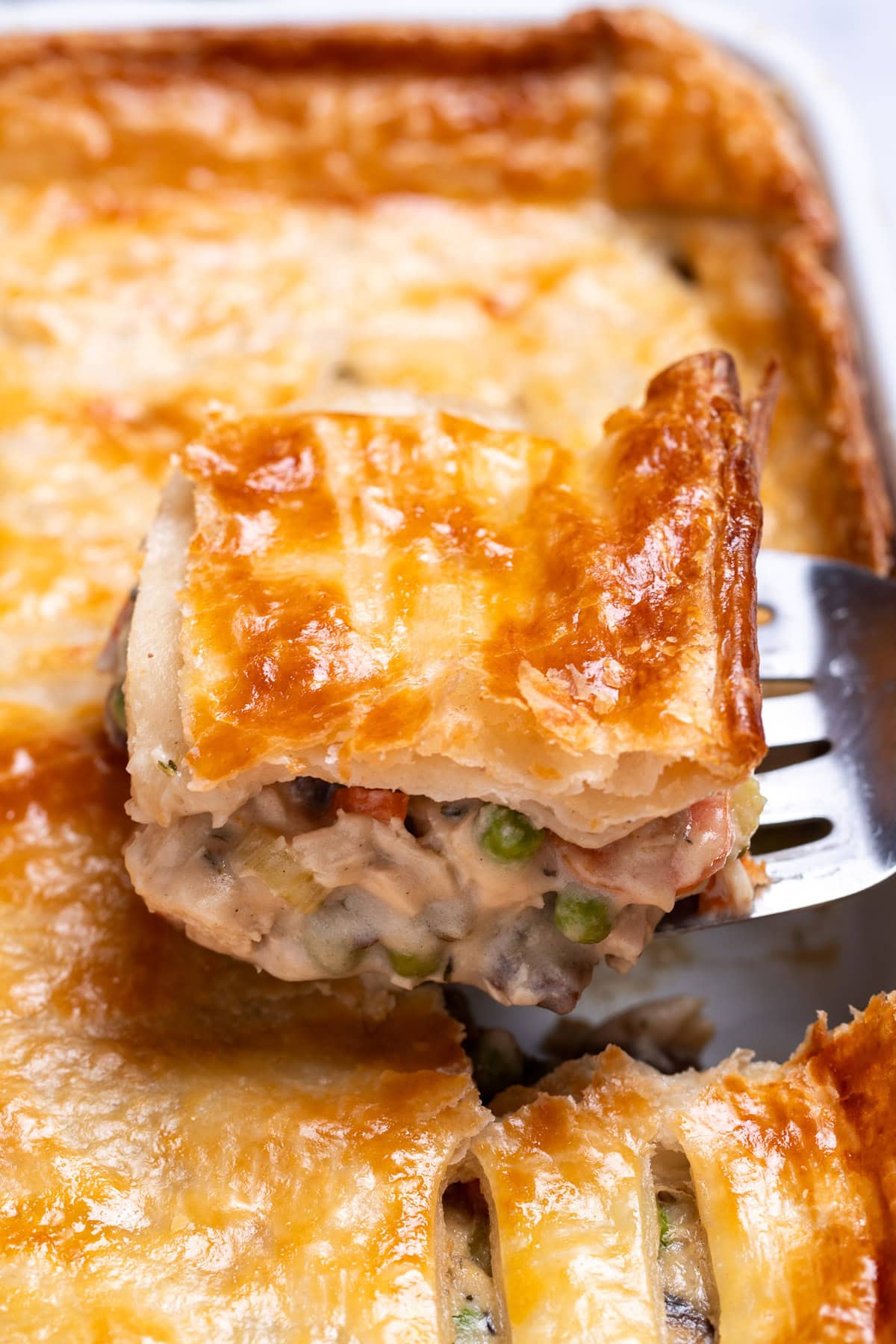A baking dish with leftover turkey pot pie casserole with a flakey puff pastry crust, and a spatula lifting some up.