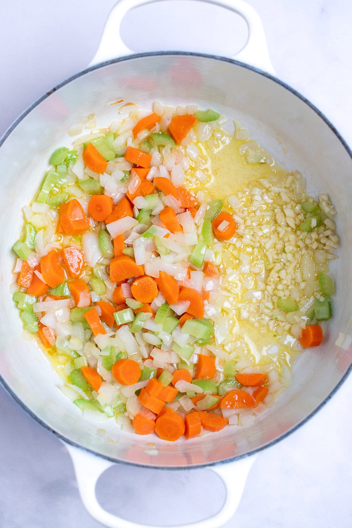 A dutch oven with sautéed carrots, onions, and celery pushed to one side, and olive oil with minced garlic on the other.