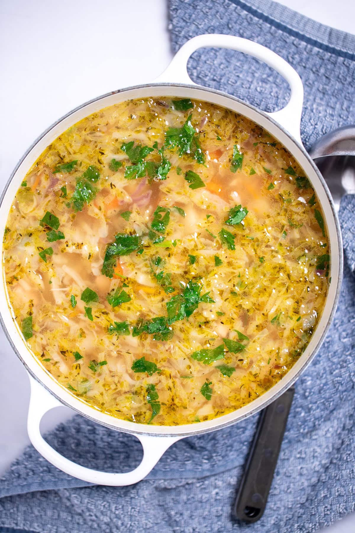 A dutch oven on a table with turkey soup topped with fresh parsley, next to a towel with a ladle on it on the table.