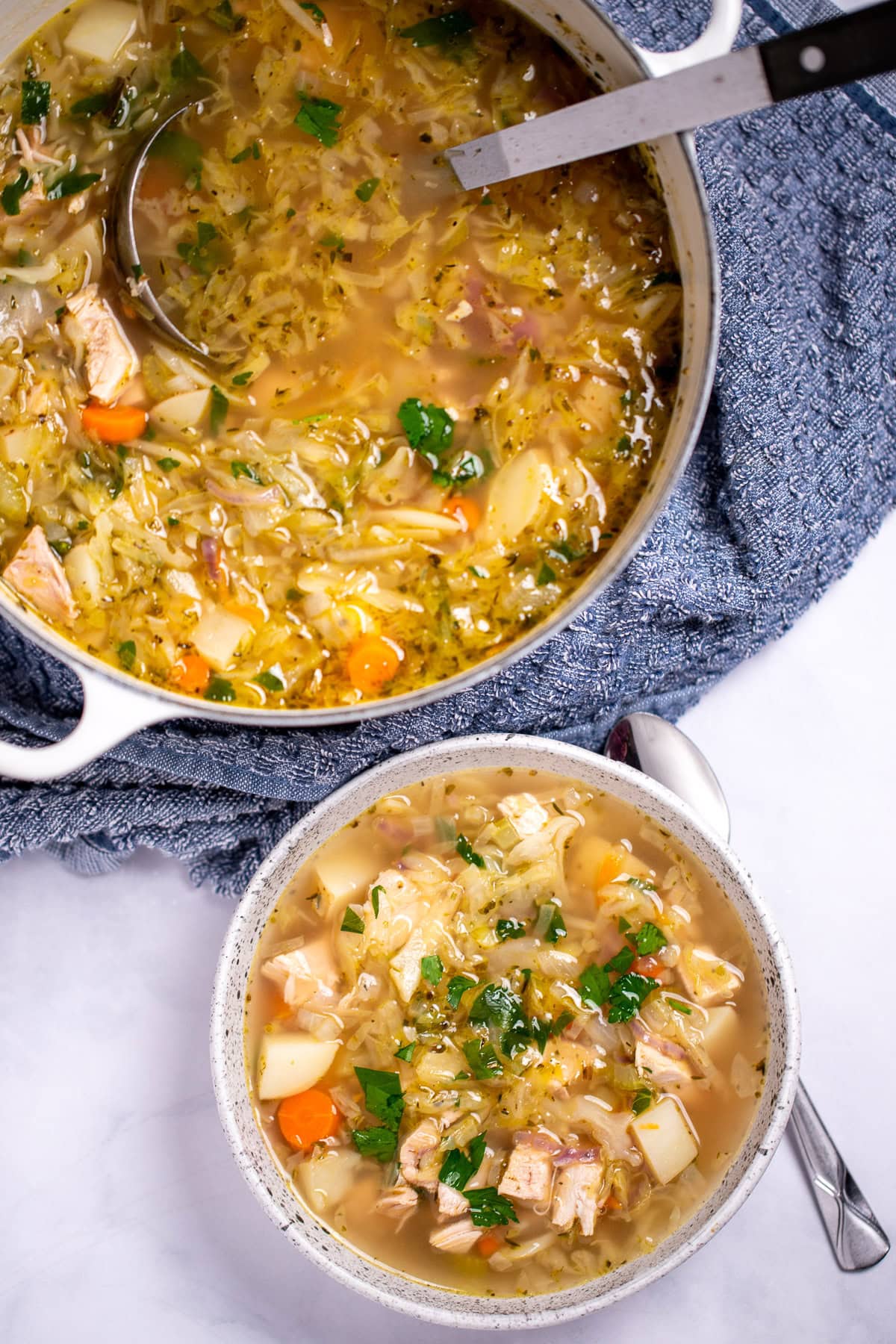 A dutch oven of leftover turkey soup with a ladle in it, and a bowl of soup topped with fresh parsley next to a spoon on the table in front of it.