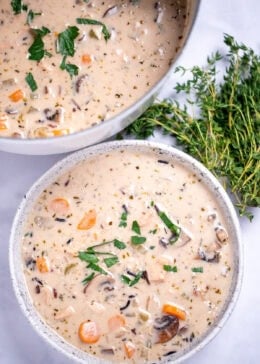 A bowl of turkey wild rice soup in front of a dutch oven of soup, with fresh thyme on the side of the bowl.