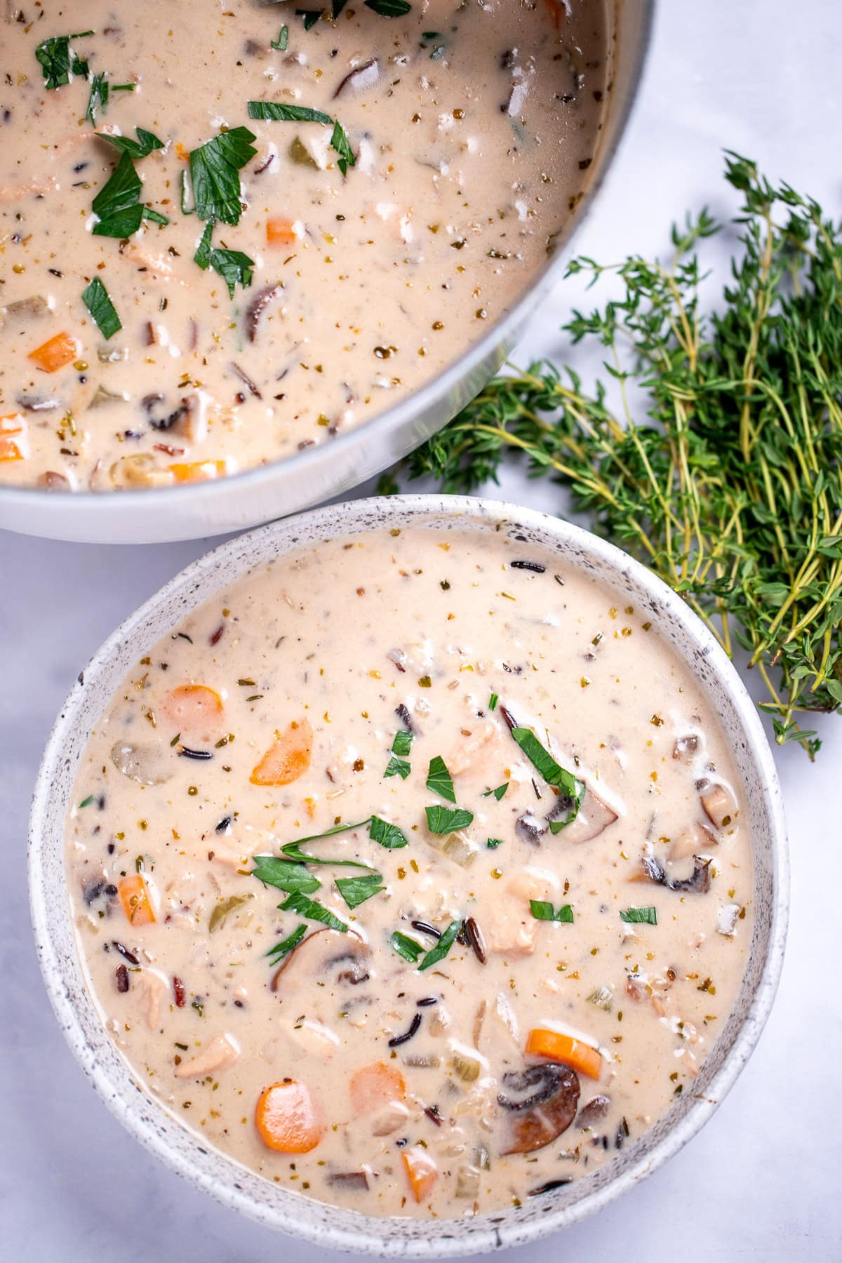 A bowl of turkey wild rice soup in front of a dutch oven of soup, with fresh thyme on the side of the bowl.