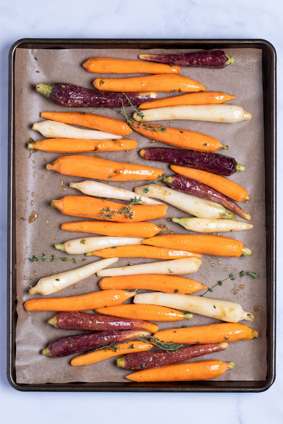A sheet pan lined with parchment paper with rainbow carrots covered in oil and spices in a single layer, with sprigs of fresh thyme, before being roasted.