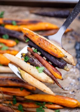 A spatula lifting roasted rainbow carrots from a sheet pan lined with parchment paper.