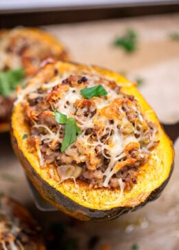 A sheet pan lined with parchment paper with four halves of acorn squash stuffed with ground sausage and vegetables and topped with melted mozzarella cheese and fresh parsley, with a spatula lifting one up.