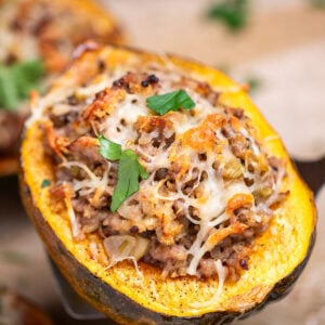 A sheet pan lined with parchment paper with four halves of acorn squash stuffed with ground sausage and vegetables and topped with melted mozzarella cheese and fresh parsley, with a spatula lifting one up.