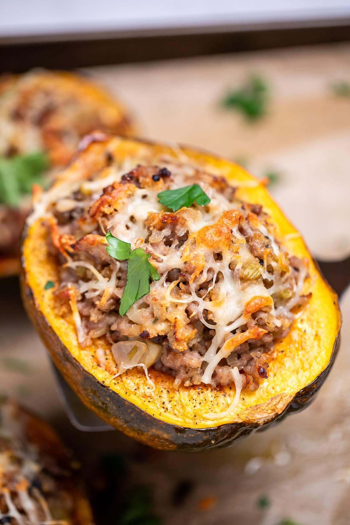 A sheet pan lined with parchment paper with four halves of acorn squash stuffed with ground sausage and vegetables and topped with melted mozzarella cheese and fresh parsley, with a spatula lifting one up.