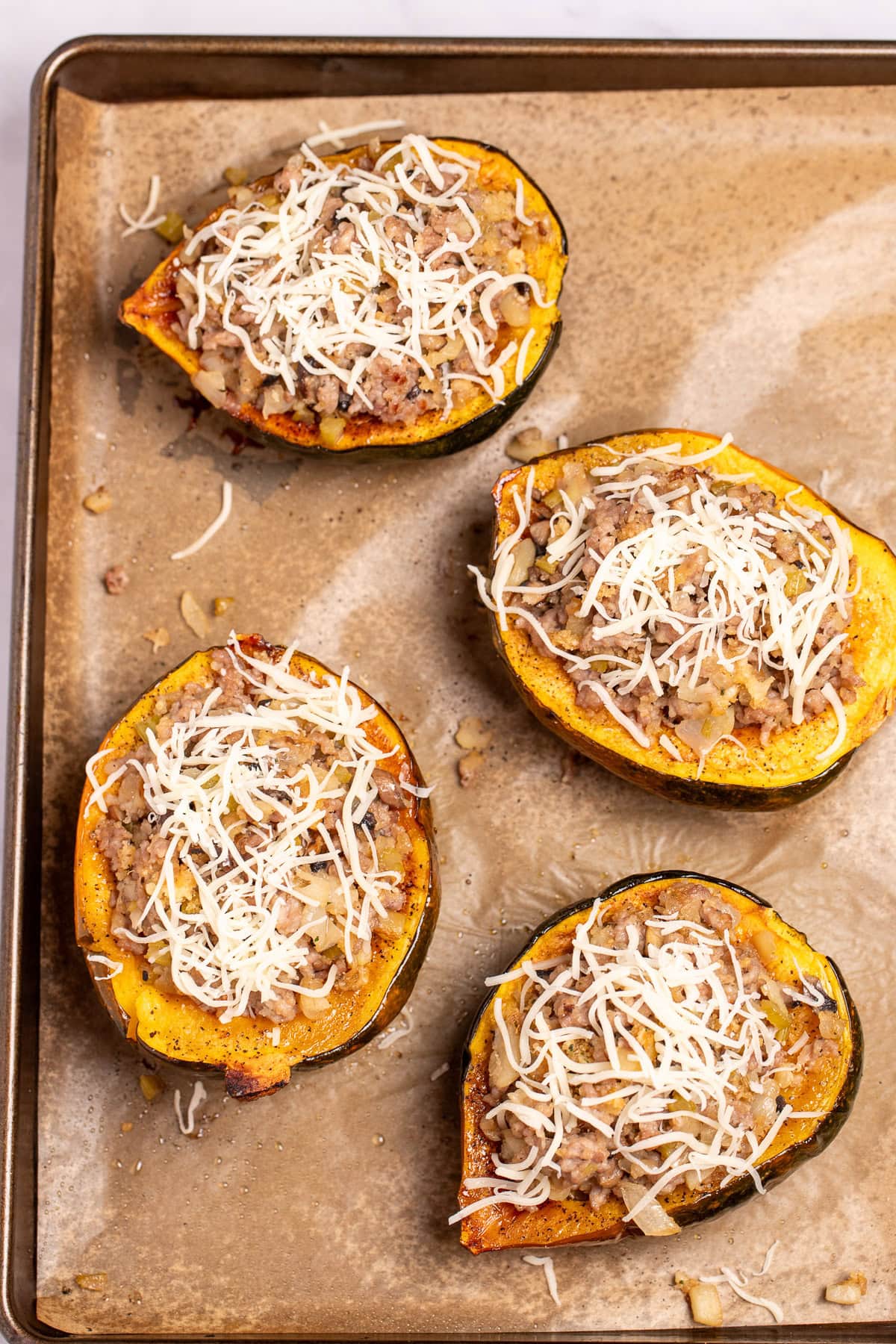 A sheet pan lined with parchment paper with four acorn squash halves full of stuffing and topped with shredded mozzarella cheese before being stuffed.