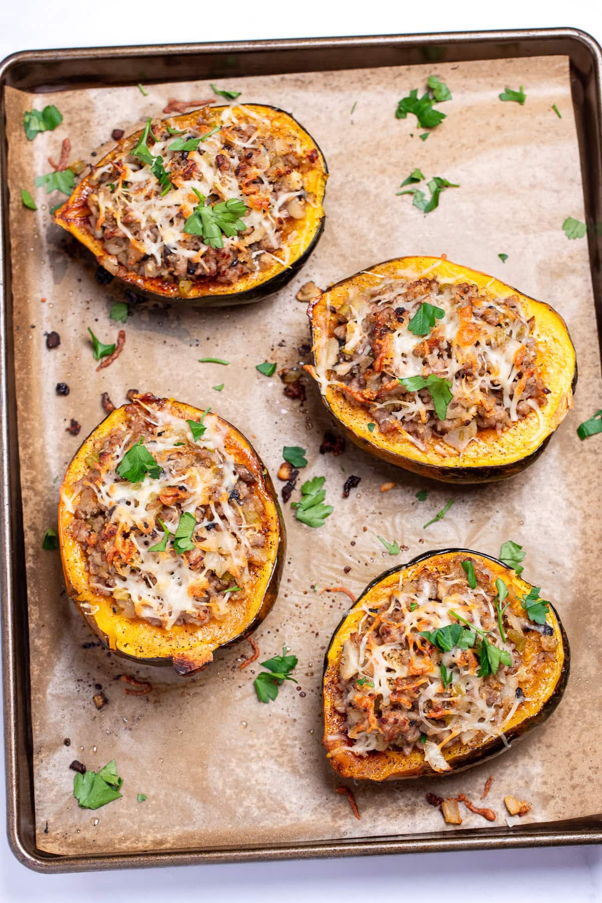 A sheet pan lined with parchment paper with four halves of acorn squash stuffed with ground sausage and vegetables and topped with melted mozzarella cheese and fresh parsley.