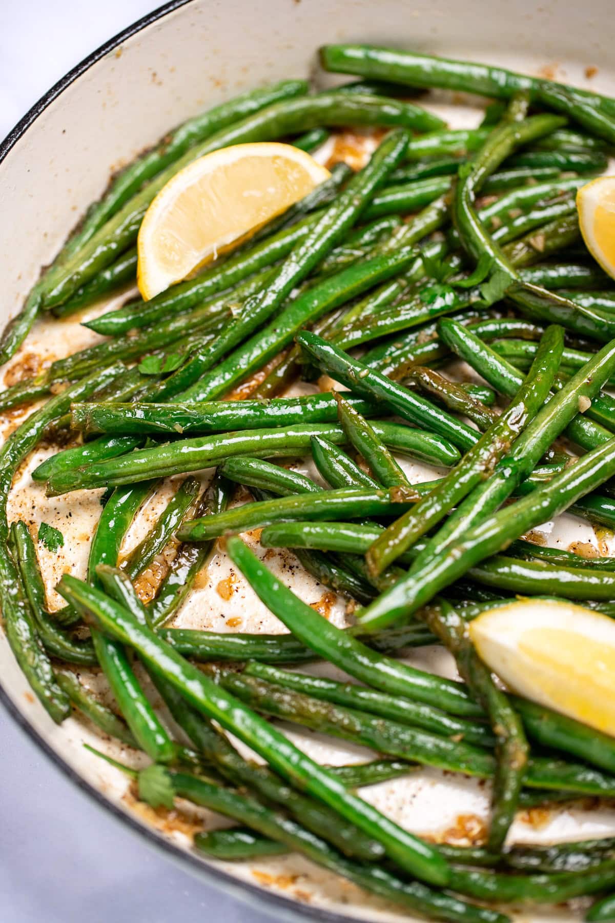 A sauté pan with cooked green beans with olive oil, salt, and pepper, and three lemon wedges resting on top.