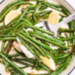 A sauté pan with cooked green beans with olive oil, salt, and pepper, and three lemon wedges resting on top, and a spatula lifting up some green beans.