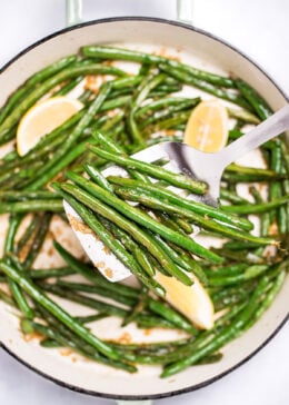 A sauté pan with cooked green beans with olive oil, salt, and pepper, and three lemon wedges resting on top, and a spatula lifting up some green beans.