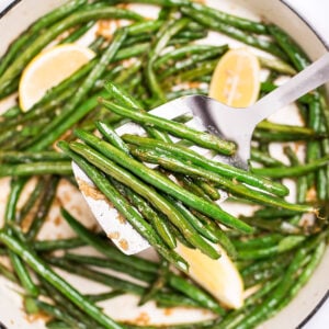 A sauté pan with cooked green beans with olive oil, salt, and pepper, and three lemon wedges resting on top, and a spatula lifting up some green beans.