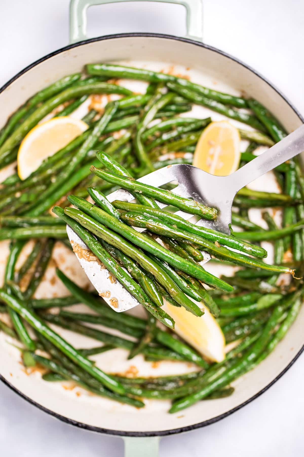 A sauté pan with cooked green beans with olive oil, salt, and pepper, and three lemon wedges resting on top, and a spatula lifting up some green beans.