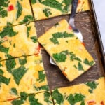 A sheet pan with fully cooked eggs with spinach and bell pepper, cut into pieces, with a spatula holding a piece and resting on the sheet pan.