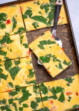 A sheet pan with fully cooked eggs with spinach and bell pepper, cut into pieces, with a spatula holding a piece and resting on the sheet pan.