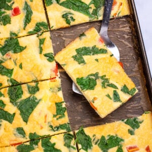 A sheet pan with fully cooked eggs with spinach and bell pepper, cut into pieces, with a spatula holding a piece and resting on the sheet pan.