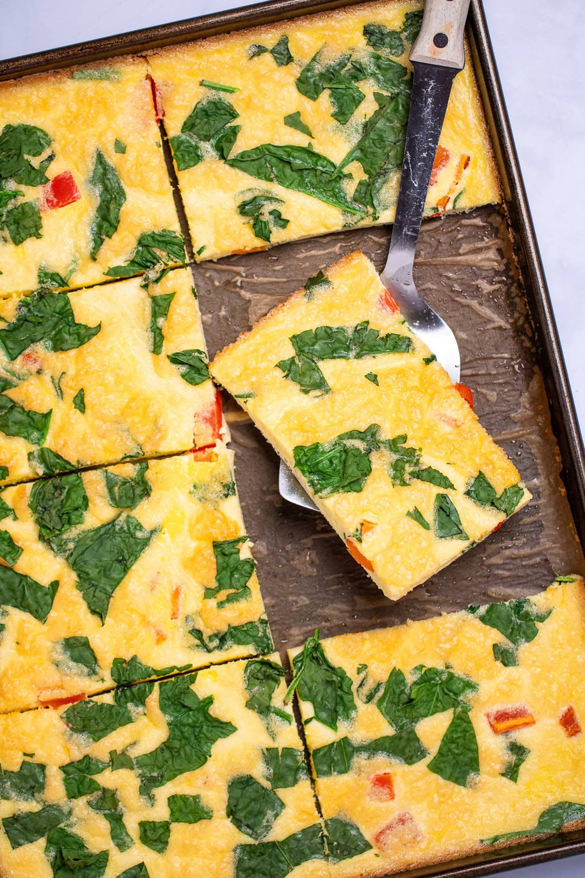 A sheet pan with fully cooked eggs with spinach and bell pepper, cut into pieces, with a spatula holding a piece and resting on the sheet pan.