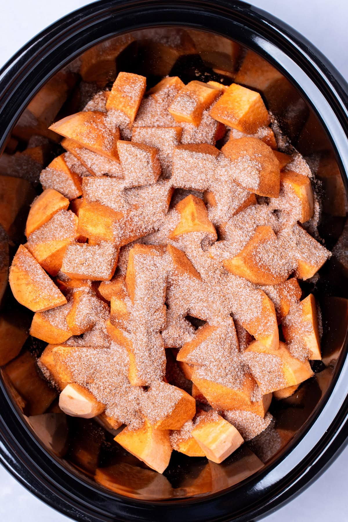 A slow cooker with sweet potato pieces topped with sugar and spices before being cooked.