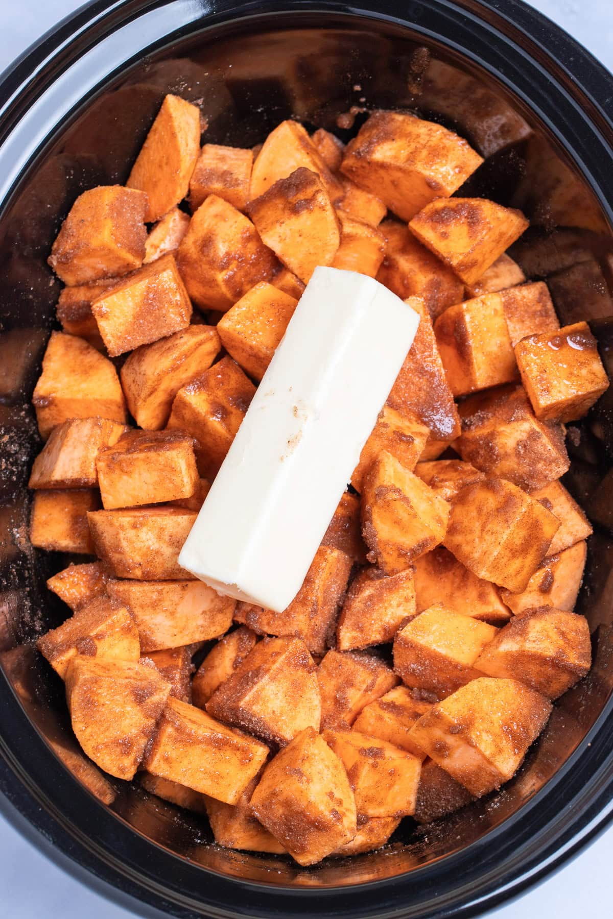 A slow cooker with sweet potato pieces stirred together with sugar, spices, and orange juice with a stick of butter on top, before being cooked.