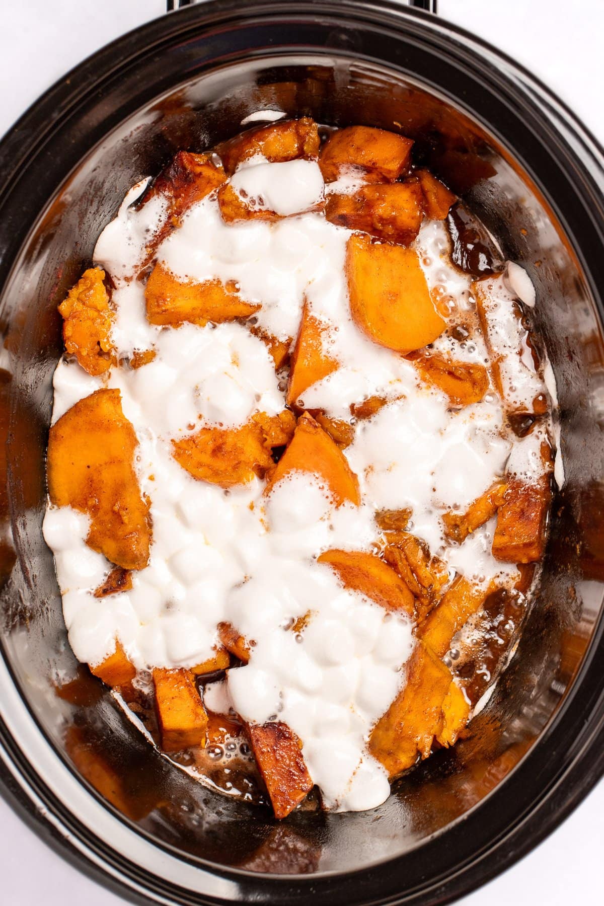 A slow cooker with candied sweet potatoes sitting in a sugar sauce with melted marshmallows on top.