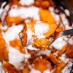 A slow cooker with candied sweet potatoes sitting in a sugar sauce with melted marshmallows on top, and a spoon lifting some up.