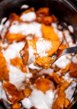 A slow cooker with candied sweet potatoes sitting in a sugar sauce with melted marshmallows on top, and a spoon lifting some up.