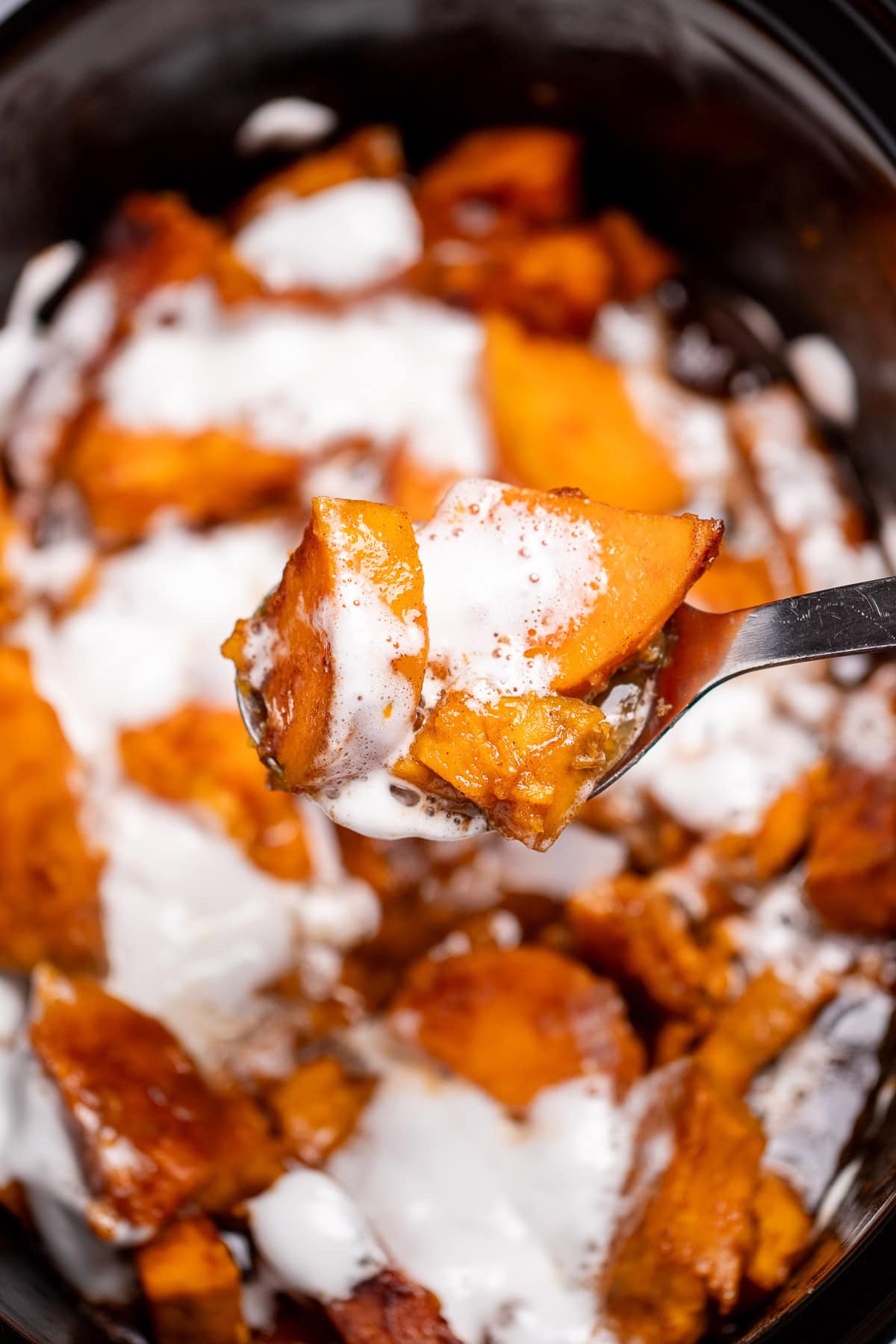A slow cooker with candied sweet potatoes sitting in a sugar sauce with melted marshmallows on top, and a spoon lifting some up.