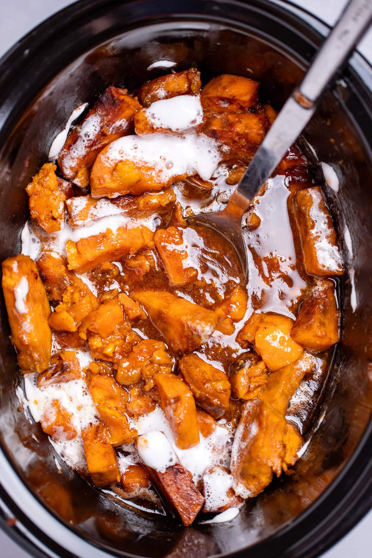 A slow cooker with candied sweet potatoes sitting in a sugar sauce with melted marshmallows on top, and a spoon resting on the side.