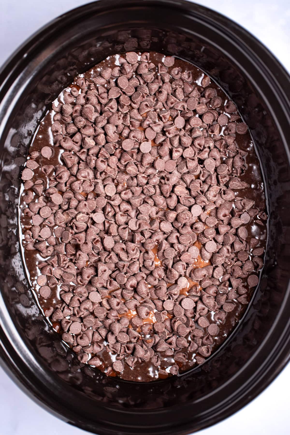 A slow cooker with cake batter topped with chocolate pudding, topped with chocolate chips before cooking.