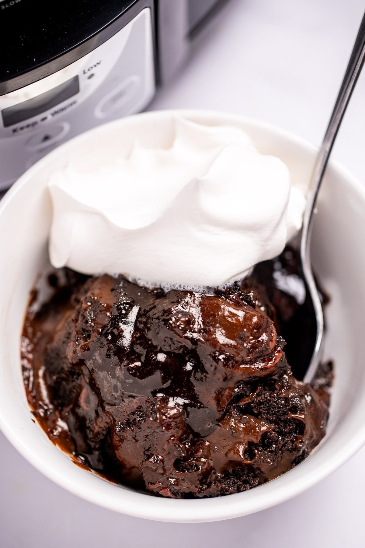 A bowl of chocolate lava cake topped with whipped cream and a spoon, in front of a slow cooker on a table.